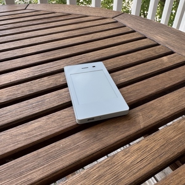 LightPhoneII.jpeg White feature phone lying face-down on a wooden slatted outdoor table, with white railings and green foliage visible in the background.